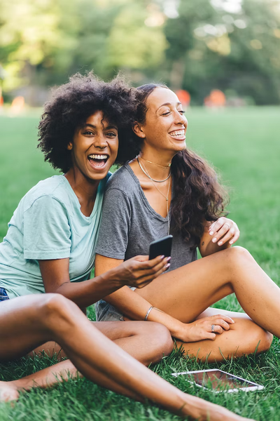 Zwei lachende Frauen sitzen nebeneinander auf einer Wiese und schauen gemeinsam auf ein Smartphone