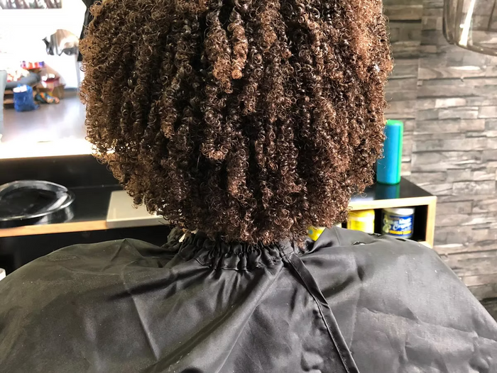 Rückenansicht einer Person mit dichten, braunen Locken, die mit einem Friseurumhang im Salon sitzt.