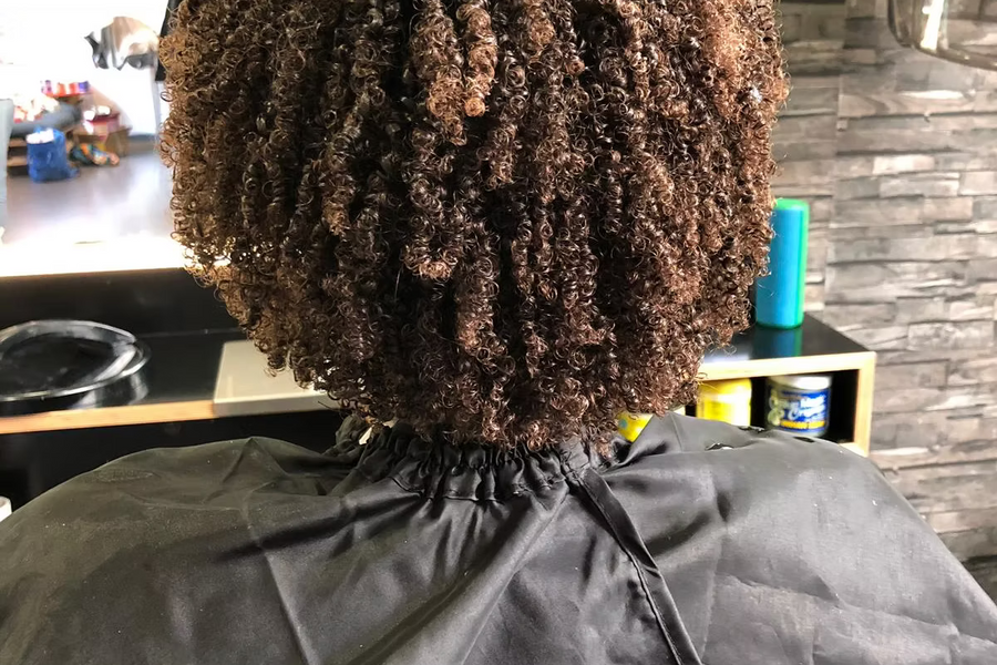 Rückenansicht einer Person mit dichten, braunen Locken, die mit einem Friseurumhang im Salon sitzt.