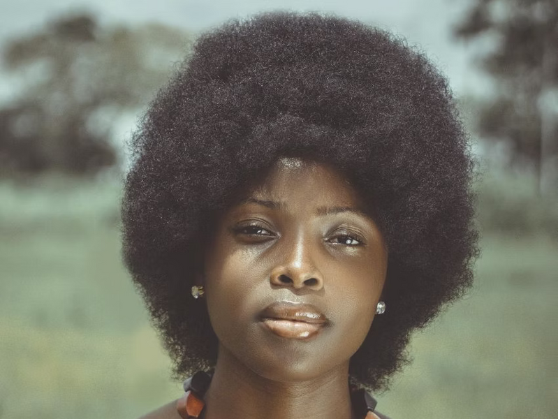 Nahaufnahme-Portrait einer Frau mit voluminösem Afro vor verschwommenem Grün im Hintergrund.
