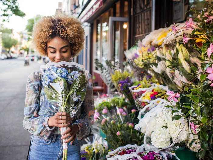 Frau mit blondem Afro-Haar riecht an einem Blumenstrauß vor einem bunten Blumenladen auf der Straße