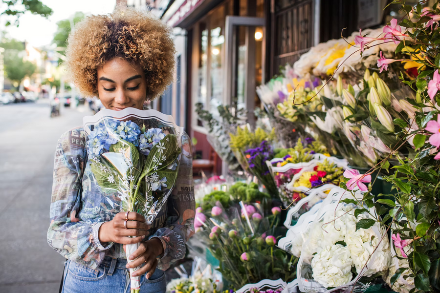 Frau mit blondem Afro-Haar riecht an einem Blumenstrauß vor einem bunten Blumenladen auf der Straße