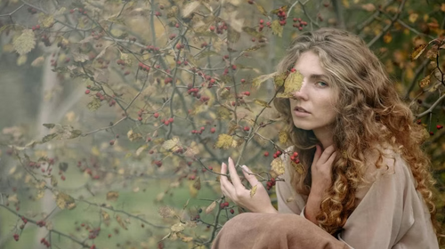 Frau mit langen, welligen Haaren sitzt zwischen herbstlichen Zweigen mit roten Beeren.