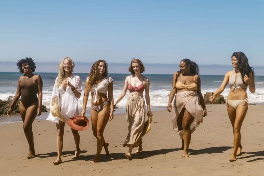 Sechs Frauen laufen lachend nebeneinander an einem sonnigen Strand