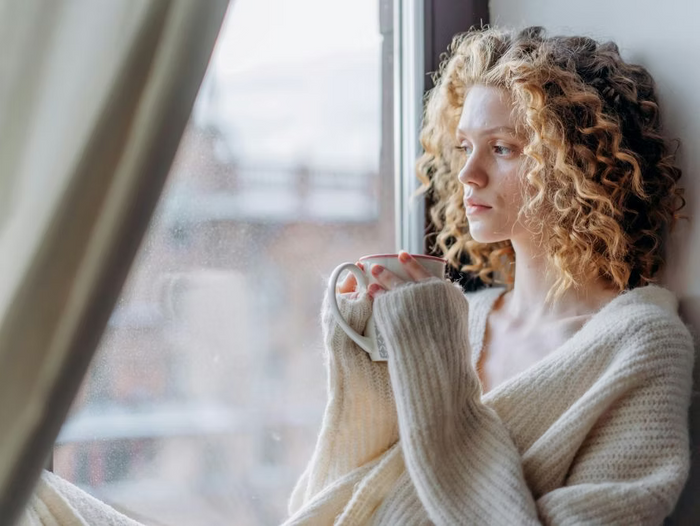 Frau mit lockigem blondem Haar sitzt mit einer Tasse in der Hand nachdenklich am Fenster und schaut hinaus.
