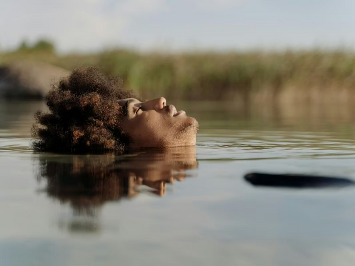 Frau mit lockigem Afro-Haar liegt mit geschlossenen Augen friedlich an der Wasseroberfläche eines Sees