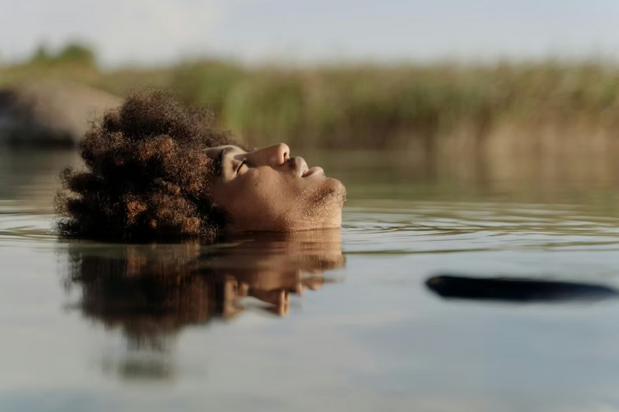 Frau mit lockigem Afro-Haar liegt mit geschlossenen Augen friedlich an der Wasseroberfläche eines Sees